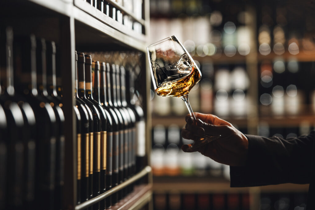 A hand holding a glass of white wine in front of a wall of wine bottles