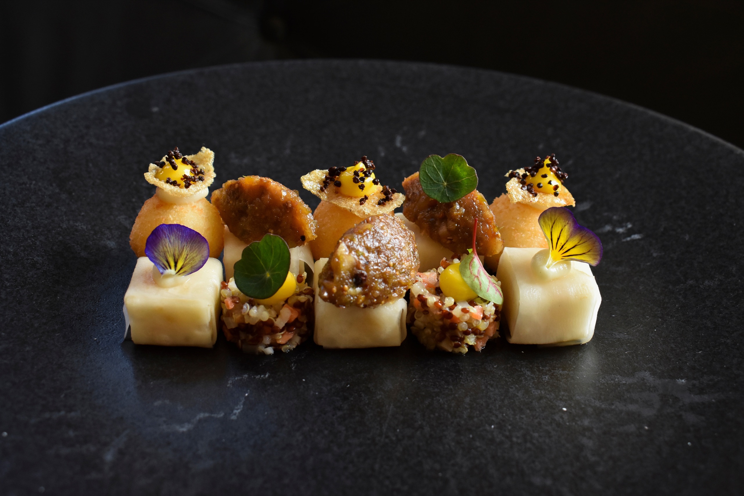Row of small canapés with edible flowers, herbs and caviar on a dark stone plate