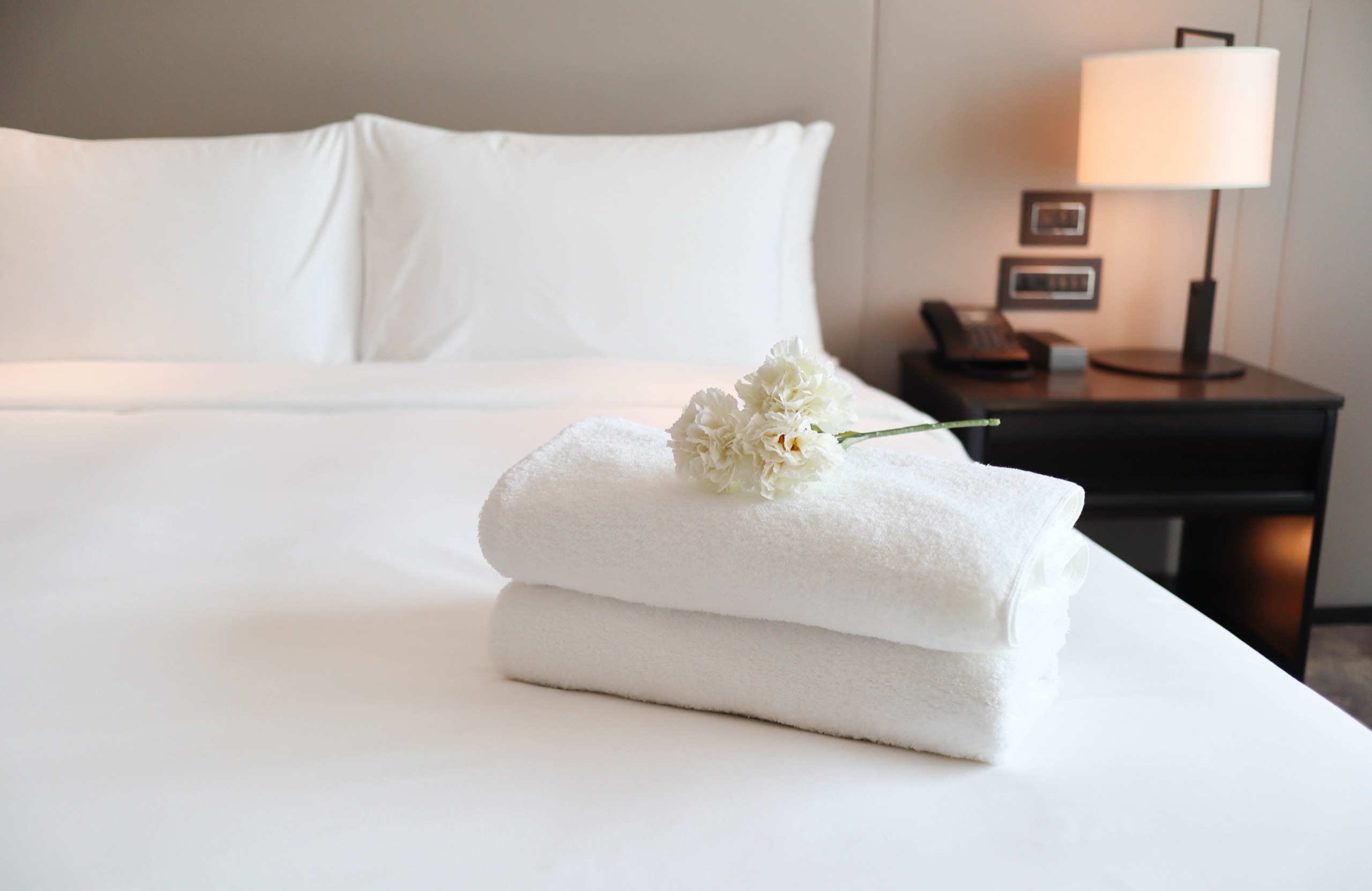 Folded white towels with a white carnation on a made bed, a bedside lamp and phone behind