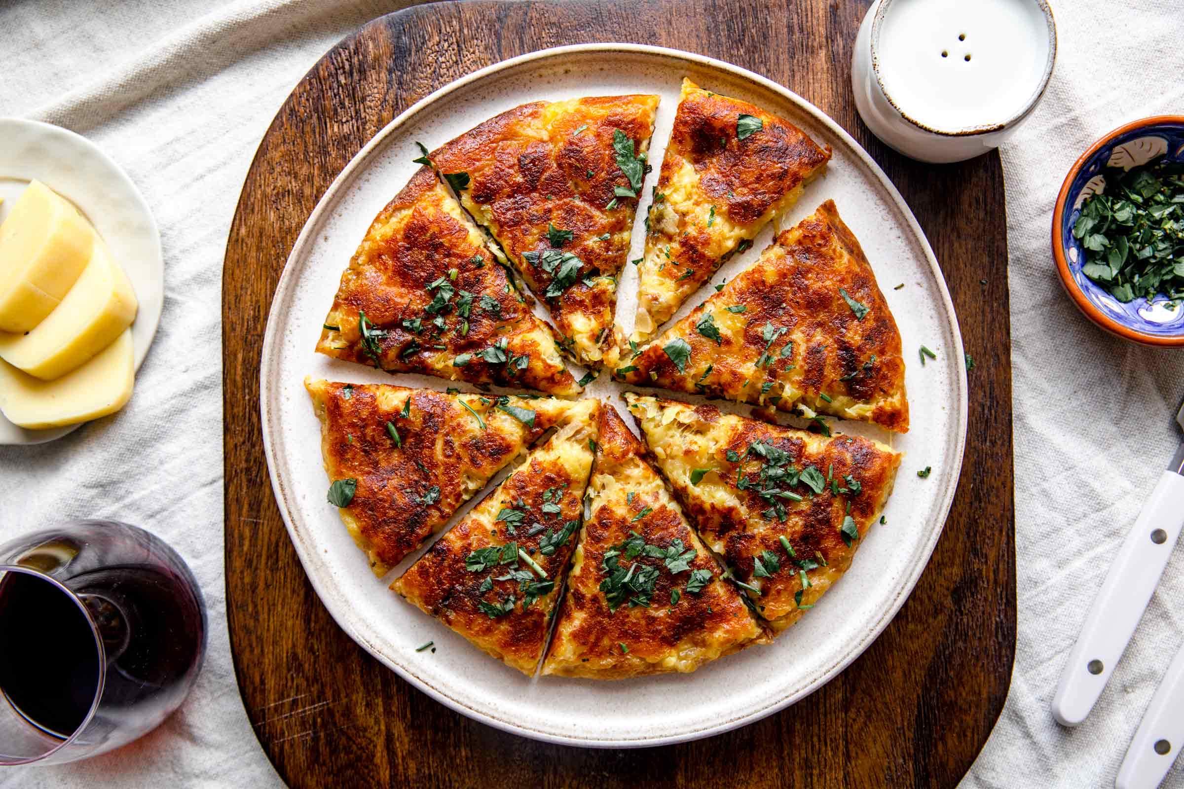 Golden potato frittata cut into wedges with chopped herbs, served on a wooden board beside a glass of red wine
